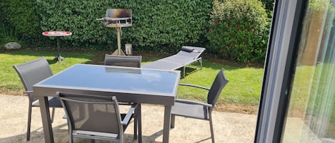 A patio with a table and chairs, a barbecue grill, and a hedge in the background.
