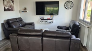 A living room with two dark brown leather sofas, a flat-screen TV mounted on the wall, a clock, and a small shelf with decorative items. - Maison Dans Lotissement Très Calme 300 Mètres du Centre Ville .10 km de la mer (La Haye)