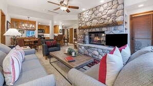 A spacious living room with a stone fireplace, a ceiling fan, and a dining area with wooden cabinets. - The Charter at Beaver Creek A260 (Beaver Creek)