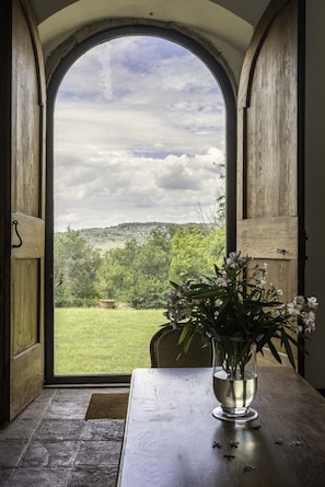 Une vue à travers une porte en arc surmontée d’un cadre en bois, qui donne sur un paysage verdoyant avec des arbres et une colline à l’horizon.