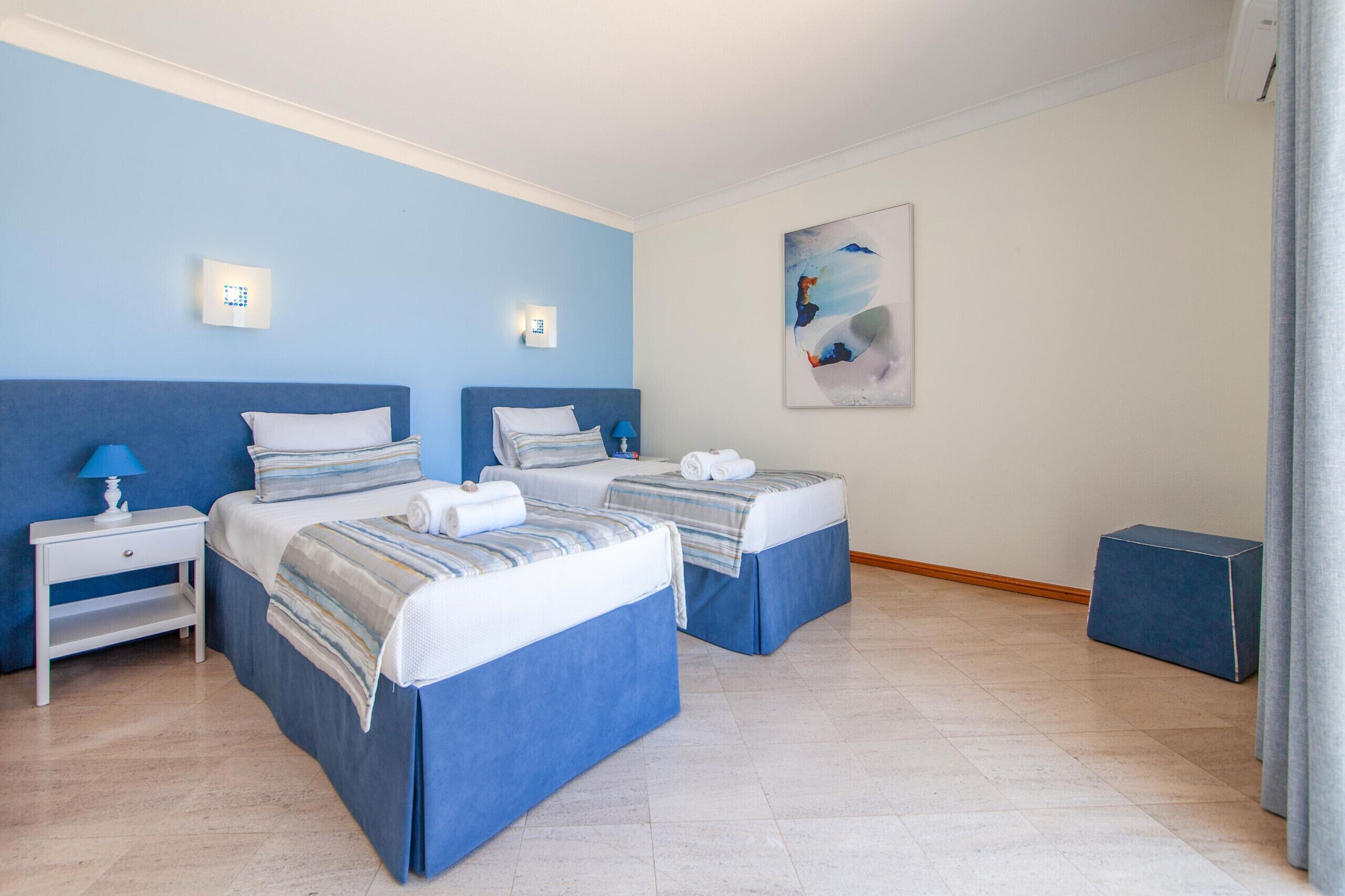 Habitación de hotel con dos camas, una mesita de noche, una lámpara, un cuadro y un pequeño otoman azul.