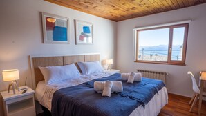 A bedroom with a wooden ceiling, a large bed with white bedding, a view of the sea through a window, and two framed artworks on the wall.