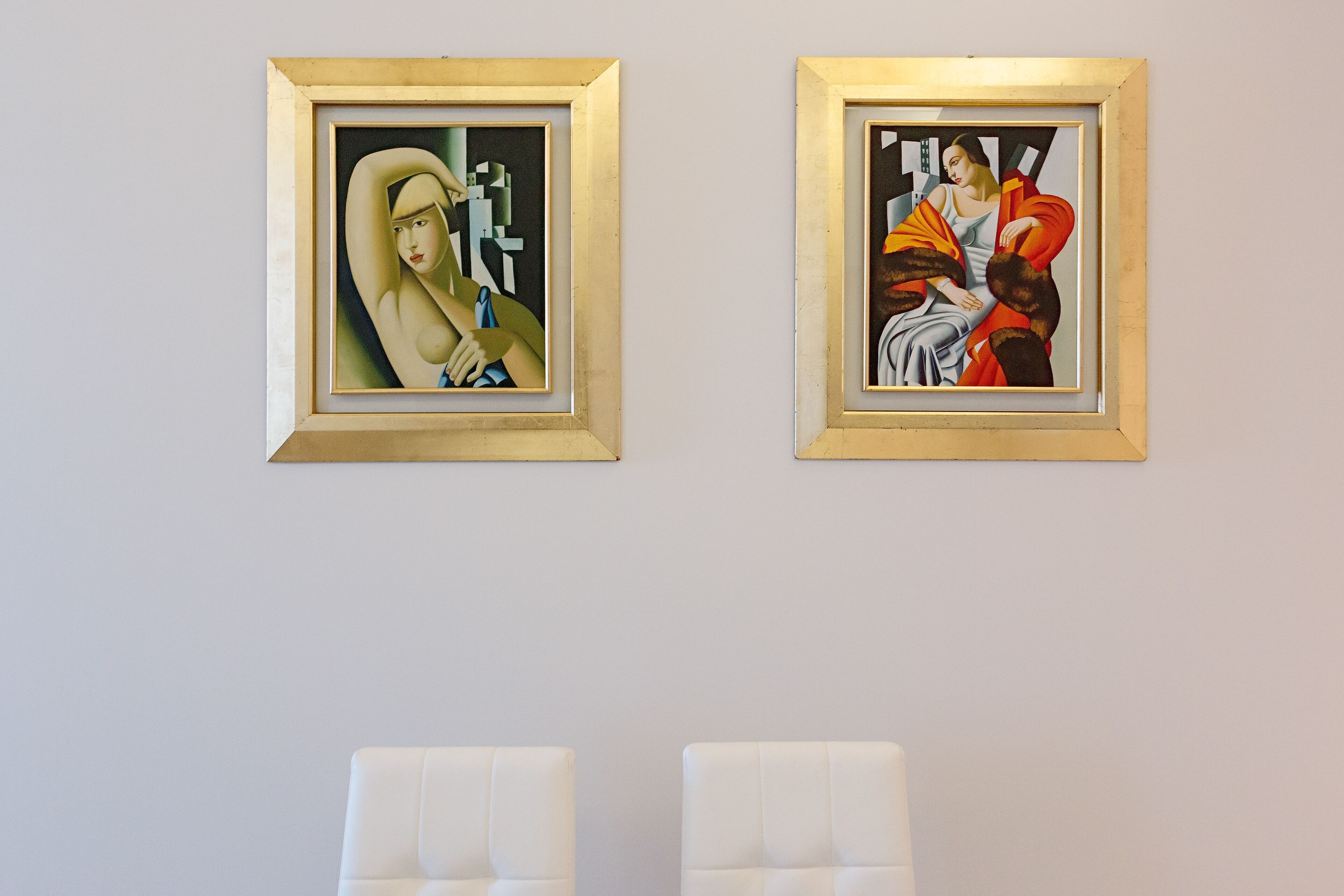 Dos obras de arte enmarcadas en una pared, con dos sillas blancas al frente.