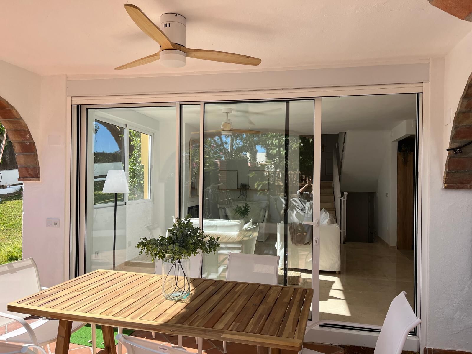 Eine Terrasse mit Holztisch und -stühlen, einem Deckenventilator und einer Glastür, die zu einem Innenraum mit Bett und Treppe führt.