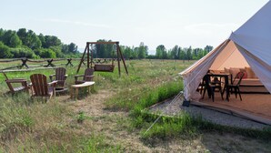 Une tente avec une table et des chaises, une balançoire et un champ d’herbe avec des arbres en arrière-plan.