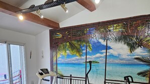 Una habitación con mesa de fútbol, equipo de ejercicio y un mural de una playa tropical.
