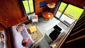 A cozy cabin interior with a wooden ceiling, a dining area with a table and chairs, a sofa with cushions, and large windows offering a view of the outdoors. - The cottage (Ploubazlanec)