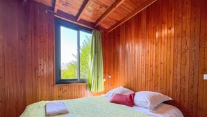 A wooden-paneled room with a bed, a window with curtains, and a nightstand with a lamp. - The cottage (Ploubazlanec)