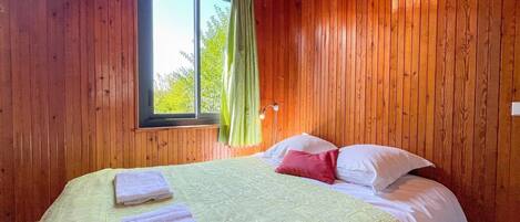A wooden-paneled room with a bed, a window with curtains, and a nightstand with a lamp.