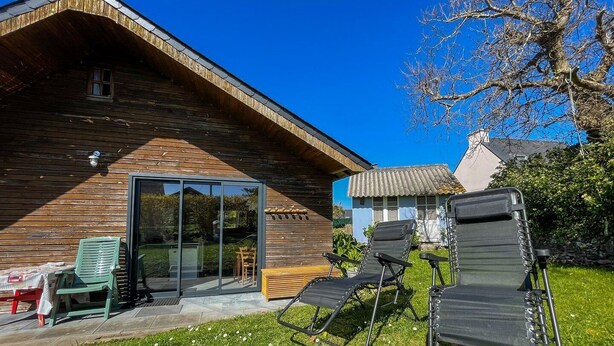 A wooden cabin with a glass door entrance, outdoor seating, and a clear blue sky. - The cottage (Ploubazlanec)