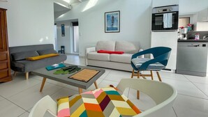 A modern living room with a grey sofa, a white sofa, a coffee table, and a colorful chair. There is a kitchen area with a dishwasher and a stove. - House Le Rohel (Pleubian)