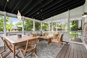 A covered outdoor patio with wooden furniture, large windows, and a view of the surrounding greenery. - Walk to the beach. Steps to the bakery. Hot Tub, King Beds, & Fire Pit. (Union Pier)