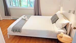 Una cama bien hecha con edredón blanco y almohadas, una mesita de noche con una vela y una ventana con cortinas.