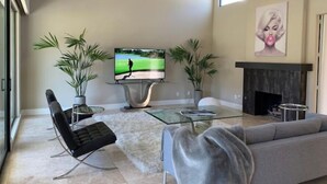 Una sala moderna con un televisor de pantalla plana, dos plantas en macetas, una mesa de centro de vidrio, un sillón de cuero negro, un sofá gris y una chimenea.