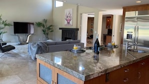 Una cocina moderna con encimera de mármol, armarios de madera y horno empotrado. Hay una zona de comedor con sofá y televisor al fondo.