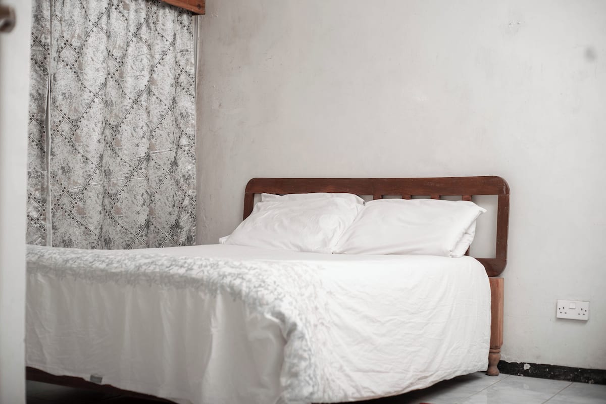 Ein Bett mit Holz-Kopfteil in einem Zimmer mit schlichten Wänden und einem gemusterten Vorhang.