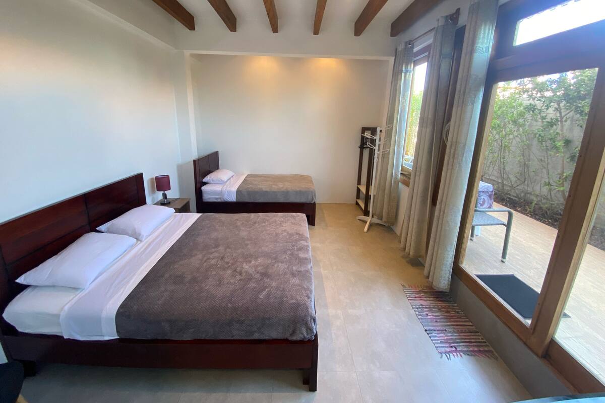 Quarto de hotel com duas camas, uma mesa de cabeceira com abajur, uma janela com cortinas e vista para o exterior.