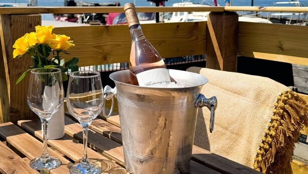 Una mesa de madera con una botella de champán, dos copas de vino y un jarrón con flores amarillas en un balcón con vistas a un puerto deportivo con veleros.