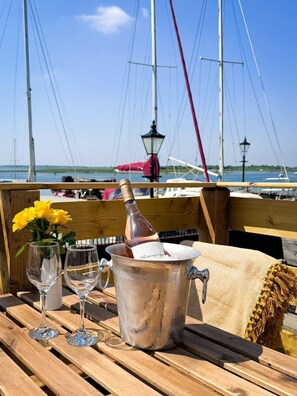 Une table en bois, sur laquelle se trouvent une bouteille de champagne, deux verres à vin et un vase de fleurs jaunes, est placée sur un balcon donnant sur une marina où sont amarrés des voiliers.