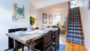 Un comedor con una mesa de madera dispuesta para cuatro, una escalera con un carpeta escocesa y un cuadro en la pared.