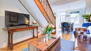Una sala de estar con una escalera de madera, un televisor de pantalla plana, una mesa de centro de madera con un jarrón de flores y un sofá de cuero.