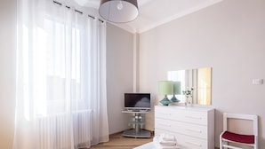 Un dormitorio con cama, televisión sobre un mueble, cómoda, silla y ventana con cortinas translúcidas.