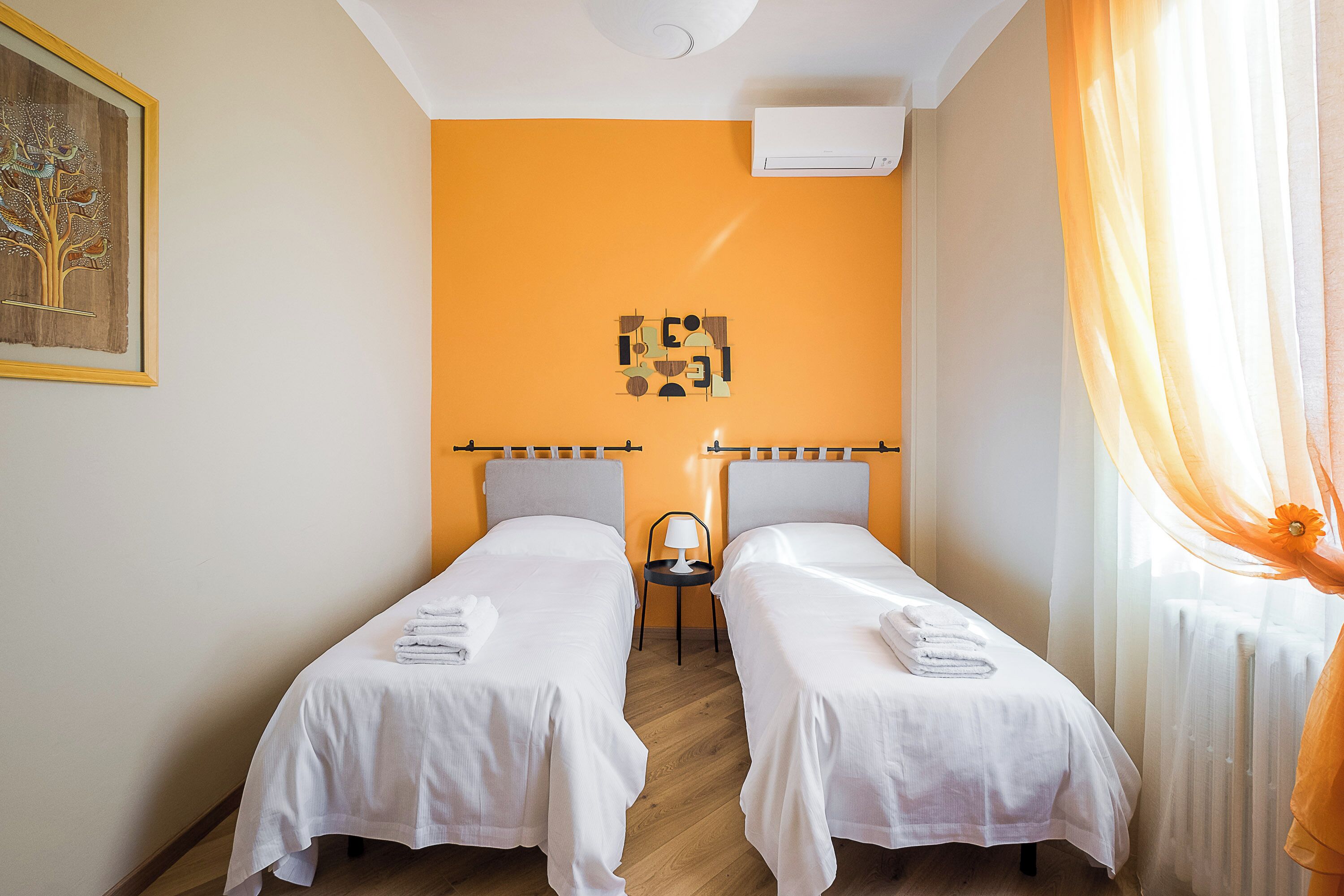 A hotel room with two single beds, a small table with a lamp, an air conditioning unit, and a window with curtains.