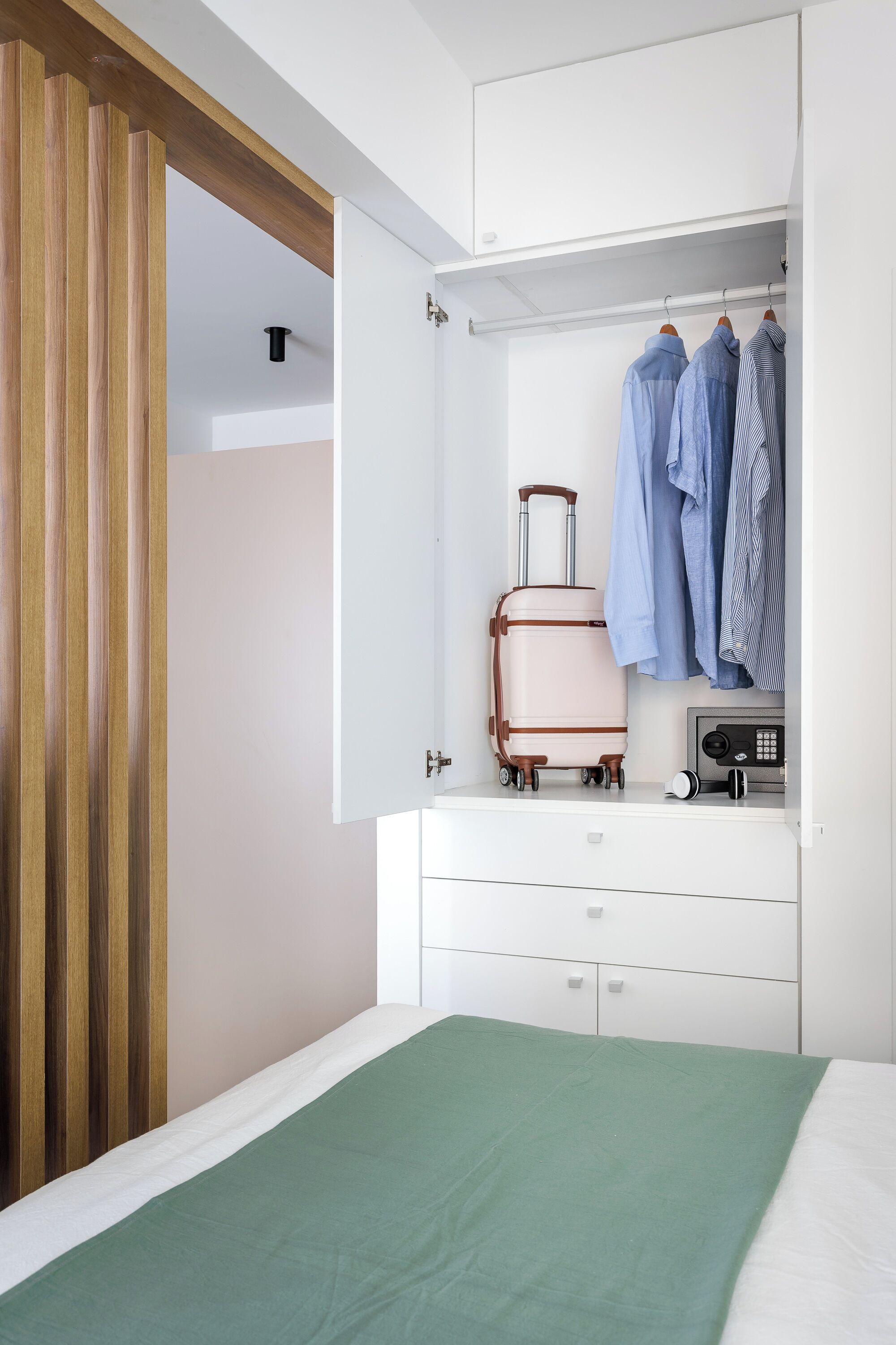 Een slaapkamer met een bed, een groene deken, een witte dressoir, een koffer en hangende kleding.