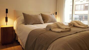 Una cama bien hecha con ropa de cama y almohadas color beige, una lámpara de noche y vista a un edificio a través de una ventana.