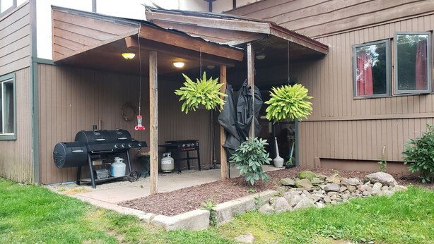 Een overdekt buitengebied met een barbecue, hangplanten en een stenen pad dat naar een huis leidt.
