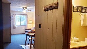 A room with a wooden wall, a ceiling fan, a window with curtains, a table with chairs, and a bathroom with a sink and towels.