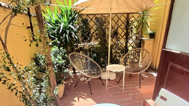 Un patio al aire libre con una mesa redonda y sillas blancas, rodeado de macetas con plantas y un paraguas grande.