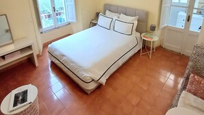 Un dormitorio con cama, mesita de noche, lámpara, un cuadro en la pared y ventana.