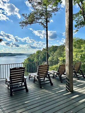 A deck with wooden chairs and tables overlooking a body of water and forested landscape.