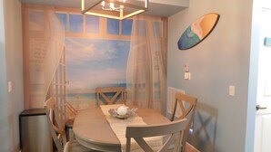 Un coin repas comprenant une table ronde, des chaises, un lustre, une fresque murale représentant une plage et une porte.