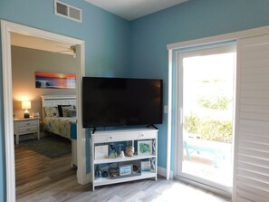 A bedroom with a TV on a white stand, a bed with a blue headboard, a nightstand with a lamp, and a sliding glass door leading to an outdoor area. - The Gardens of Beachwalk 109 (Fort Myers)