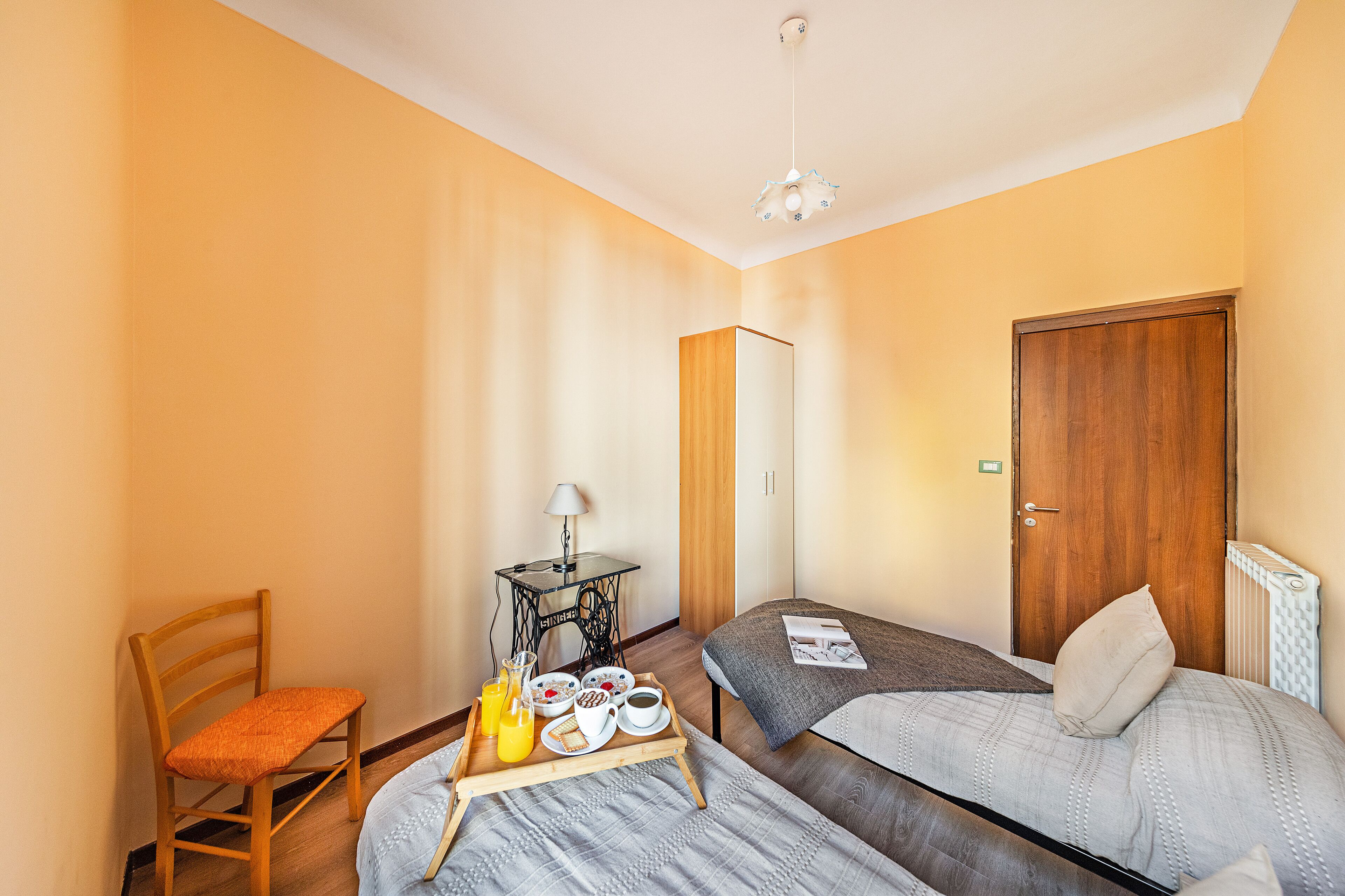 A room with two beds, a wooden chair, a small table with a lamp, and a tray with breakfast items.