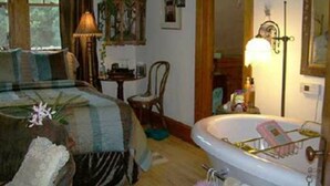 A bedroom with a bed, a chair, a desk, a lamp, a mirror, and a bathtub. - Relax in our English Garden King Suite at The Cobblestone Bed & Breakfast (Birchwood)