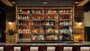 Una zona de bar con varias estanterías llenas de botellas de licores, un piano y cuatro taburetes rojos.
