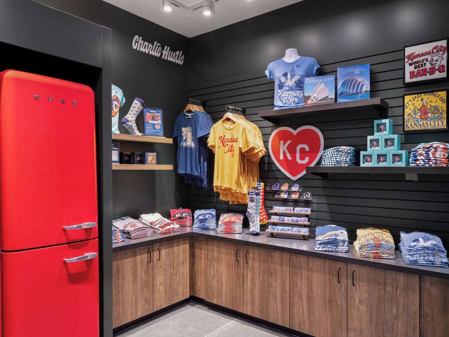 A retail space with a red refrigerator, shelves with Kansas City-themed merchandise, and a wall sign reading "Charlie Hustle".
