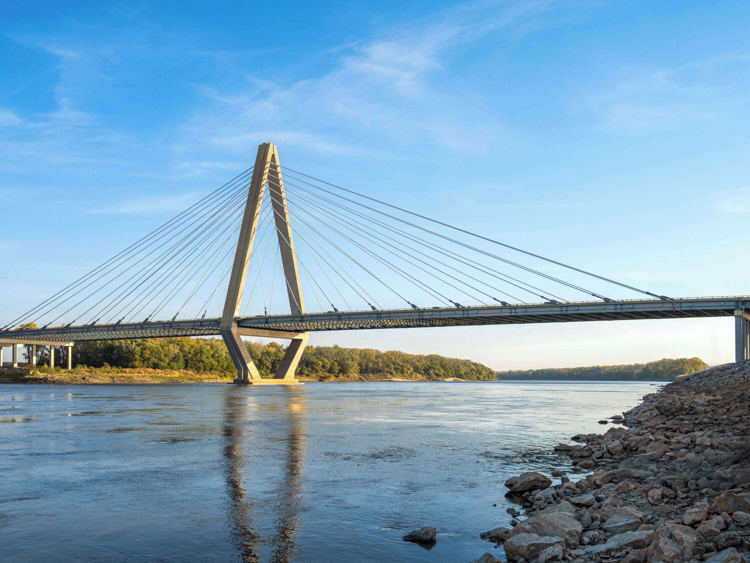 Eine Schrägseilbrücke mit einem zentralen Turm, die einen Fluss mit steinigen Ufern und Bäumen im Hintergrund überspannt.