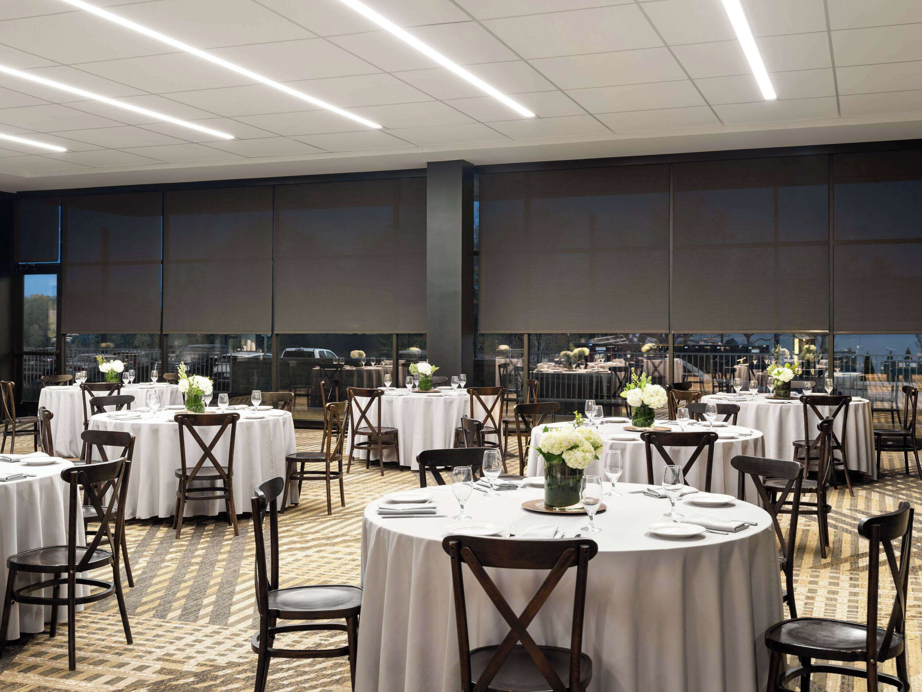 A banquet hall with round tables, white tablecloths, and dark wooden chairs.