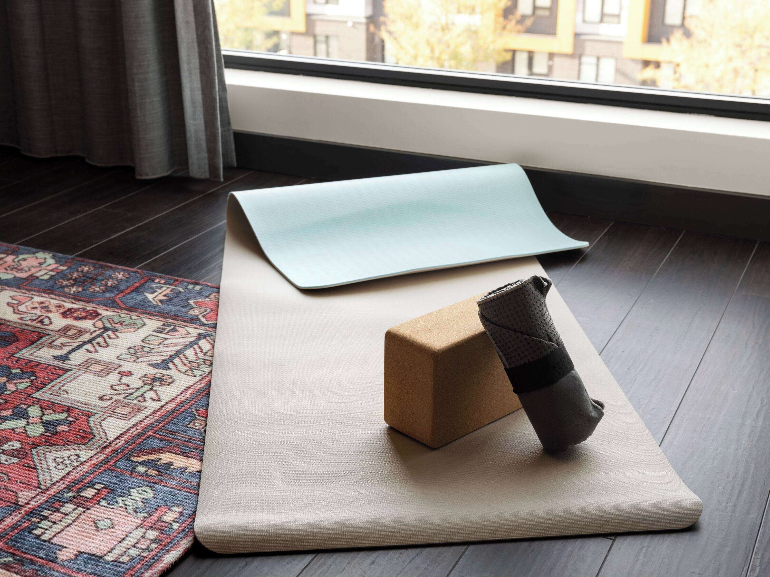 Een yogamat, een opgevouwen mat en een opgerolde mat op een houten vloer met een kamerkleed met een patroon.