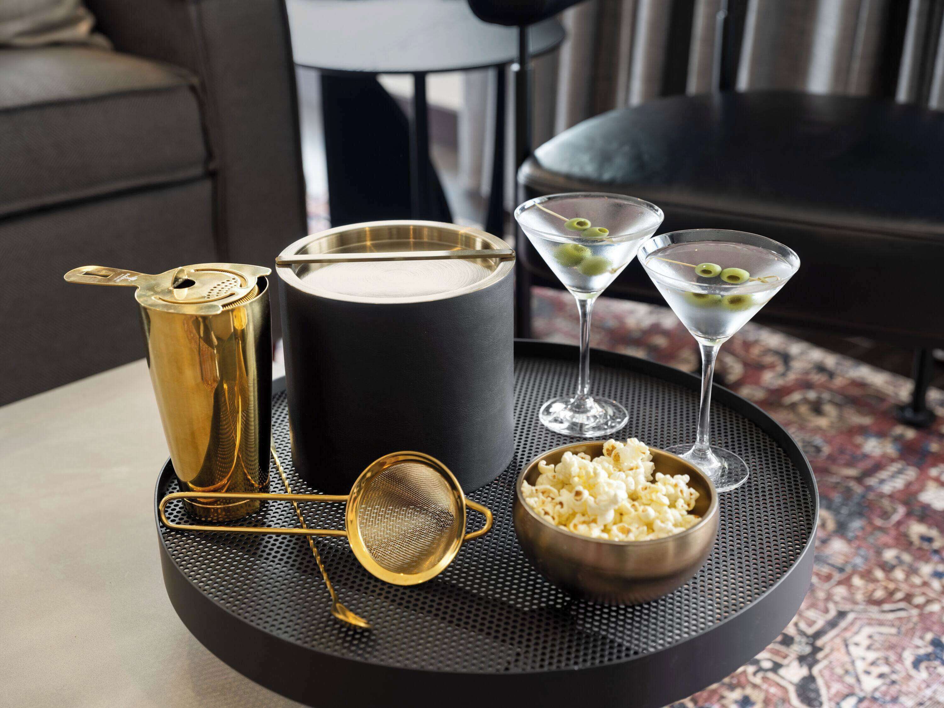 A tray with a gold cocktail shaker, a black container, two martini glasses with olives, a gold strainer, and a bowl of popcorn.
