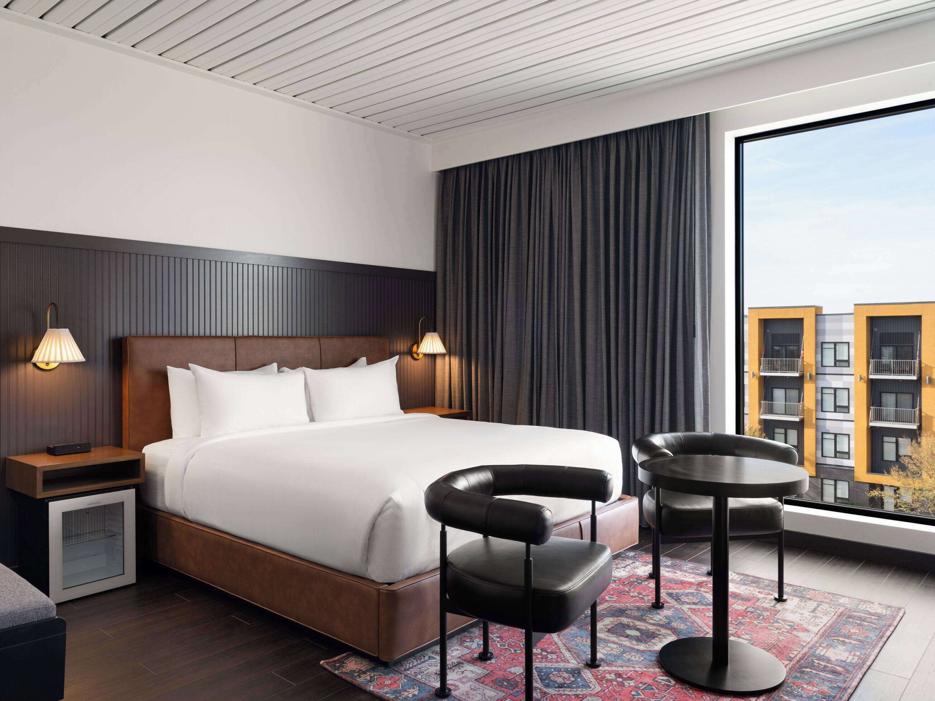 Una habitación de hotel moderna con una cama grande, dos lámparas de noche, una mesita con una silla y un ventanal con vistas a la ciudad.