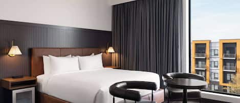 Una habitación de hotel moderna con una cama grande, dos lámparas de noche, una mesita con una silla y un ventanal con vistas a la ciudad.