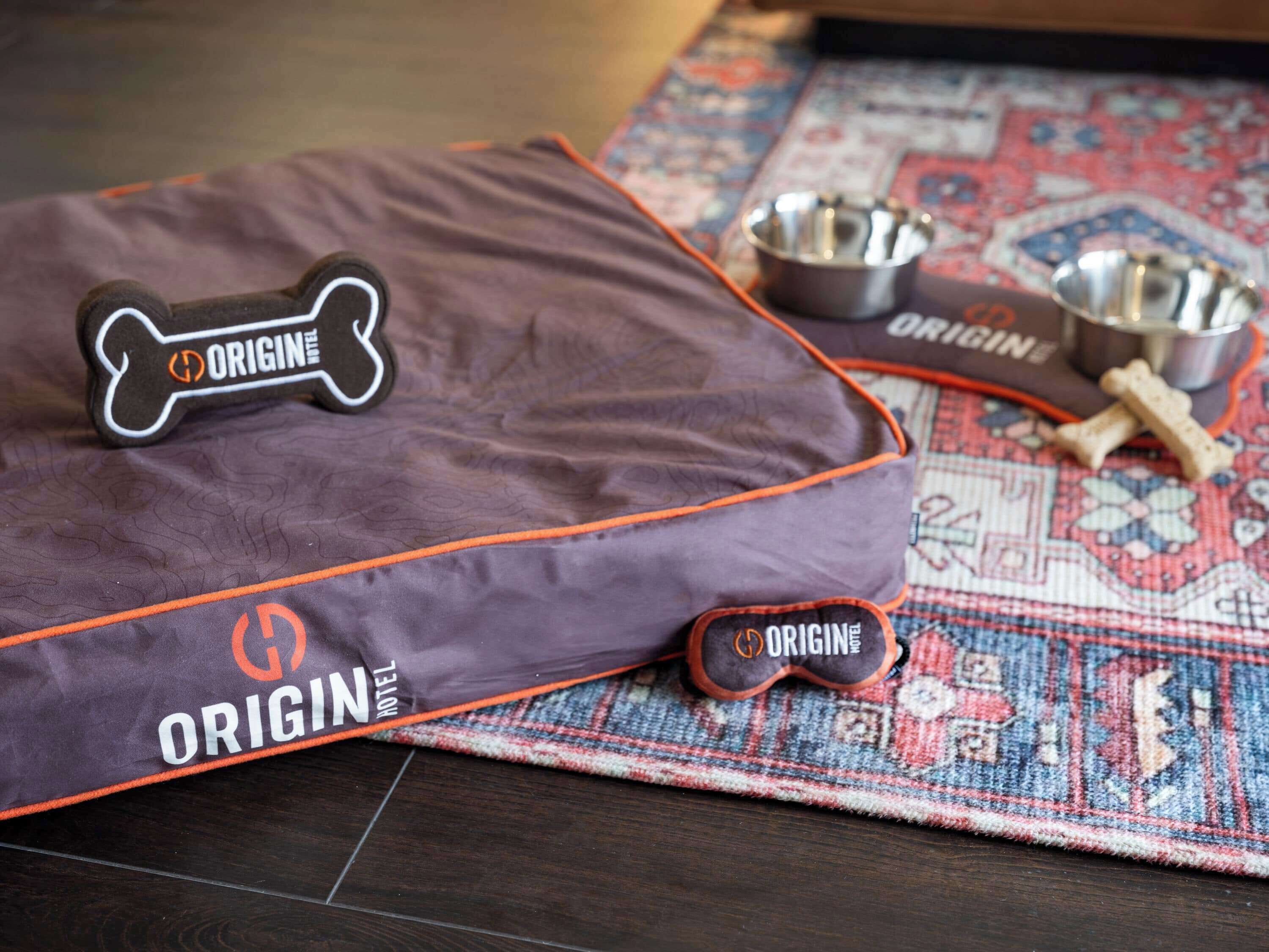 Un panier pour animaux de compagnie étiqueté « ORIGIN HOTEL », avec un coussin en forme d’os, deux gamelles en métal et deux jouets à mâcher en forme d’os, le tout posé sur un tapis à motifs.