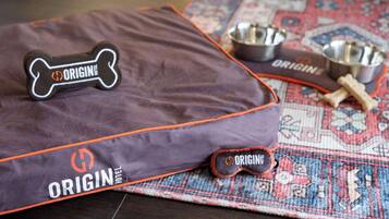 Un panier pour animaux de compagnie étiqueté « ORIGIN HOTEL », avec un coussin en forme d’os, deux gamelles en métal et deux jouets à mâcher en forme d’os, le tout posé sur un tapis à motifs.