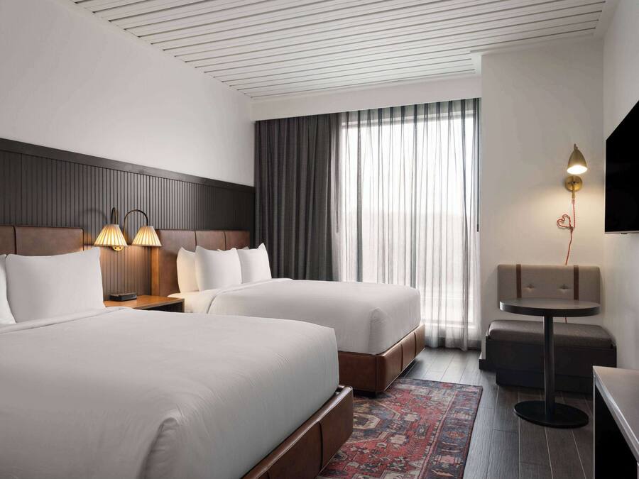 A hotel room with two beds, a wooden headboard, a nightstand with lamps, a small table, and a TV mounted on the wall.