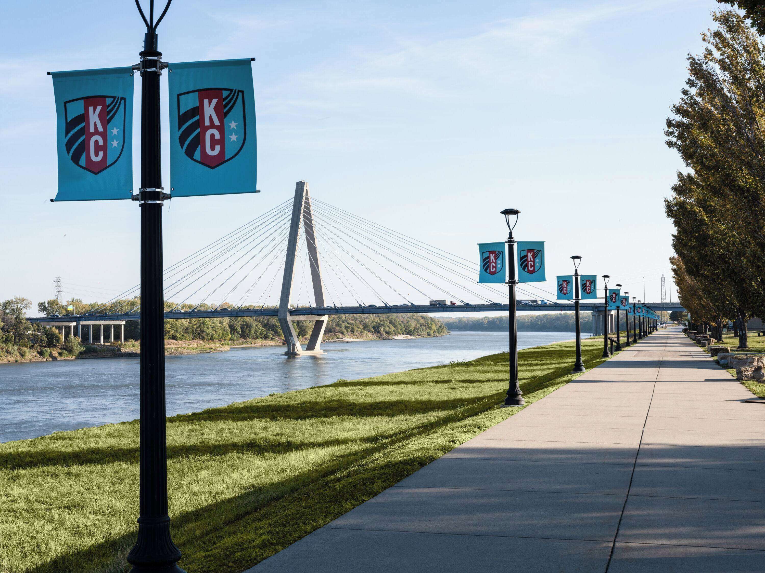 A pedestrian walkway with a river, a bridge, and street lamps with KC logos.